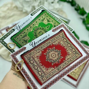 May include: Miniature decorative rug-shaped pouches in red and green, with intricate gold patterns. Each pouch is displayed on a white card with a decorative border and the text "Yasmina's Henna Night". The red pouch has "December 2025" printed on the card.