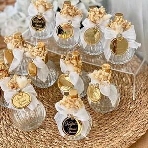 Nine small, clear, ribbed glass perfume bottles with gold tops, white bows, dried flowers, and gold tags are arranged on a woven mat and clear stand.