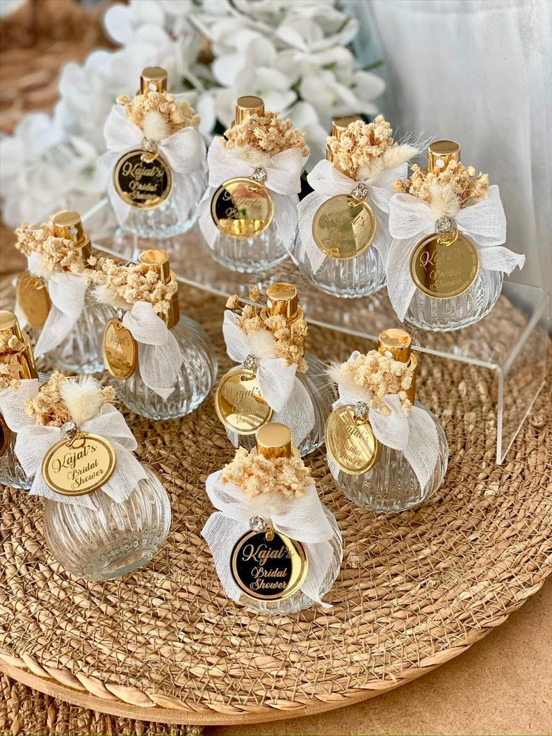 Nine small, clear, ribbed glass perfume bottles with gold tops, white bows, dried flowers, and gold tags are arranged on a woven mat and clear stand.