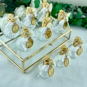 Eight small, clear, ribbed glass perfume bottles with gold tops, white bows, dried flowers, and gold tags are arranged on a gold-framed glass tray.