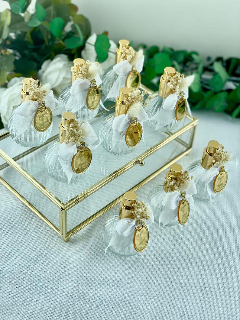 Eight small, clear, ribbed glass perfume bottles with gold tops, white bows, dried flowers, and gold tags are arranged on a gold-framed glass tray.