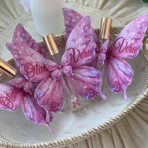 May include: A collection of small, clear glass perfume bottles with gold-colored caps, each adorned with a pink butterfly wing design. The wings feature glitter accents and the names "Olivia" and "Vera" in script. The bottles are arranged in a decorative, woven basket.