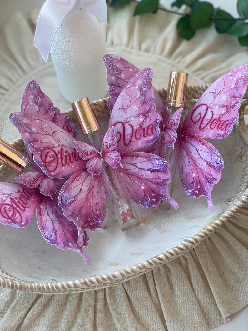 May include: A collection of small, clear glass perfume bottles with gold-colored caps, each adorned with a pink butterfly wing design. The wings feature glitter accents and the names "Olivia" and "Vera" in script. The bottles are arranged in a decorative, woven basket.
