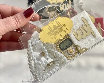 May include: A clear acrylic gift box containing a white pearl prayer bead, a gold-colored digital counter, a small white towel with gold Arabic calligraphy, and a gold-colored Ramadan Kareem tag.