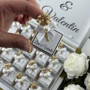 May include: A white box with a gold trim filled with small white boxes with a white bow and dried flowers. The boxes are labeled "Love & Valentin" and the lid of the box has the names "Leva & Valentin" written in black.