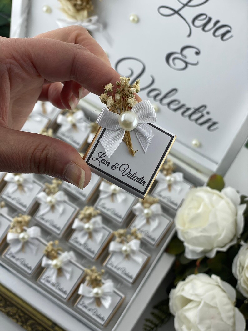 May include: A white box with a gold trim filled with small white boxes with a white bow and dried flowers. The boxes are labeled "Love & Valentin" and the lid of the box has the names "Leva & Valentin" written in black.