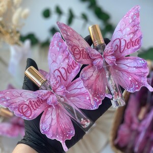 May include: Pink butterfly-shaped perfume bottles with gold caps. The butterfly wings are decorated with glitter and the names "Olivia" and "Vera". The bottles are held in a black gloved hand.