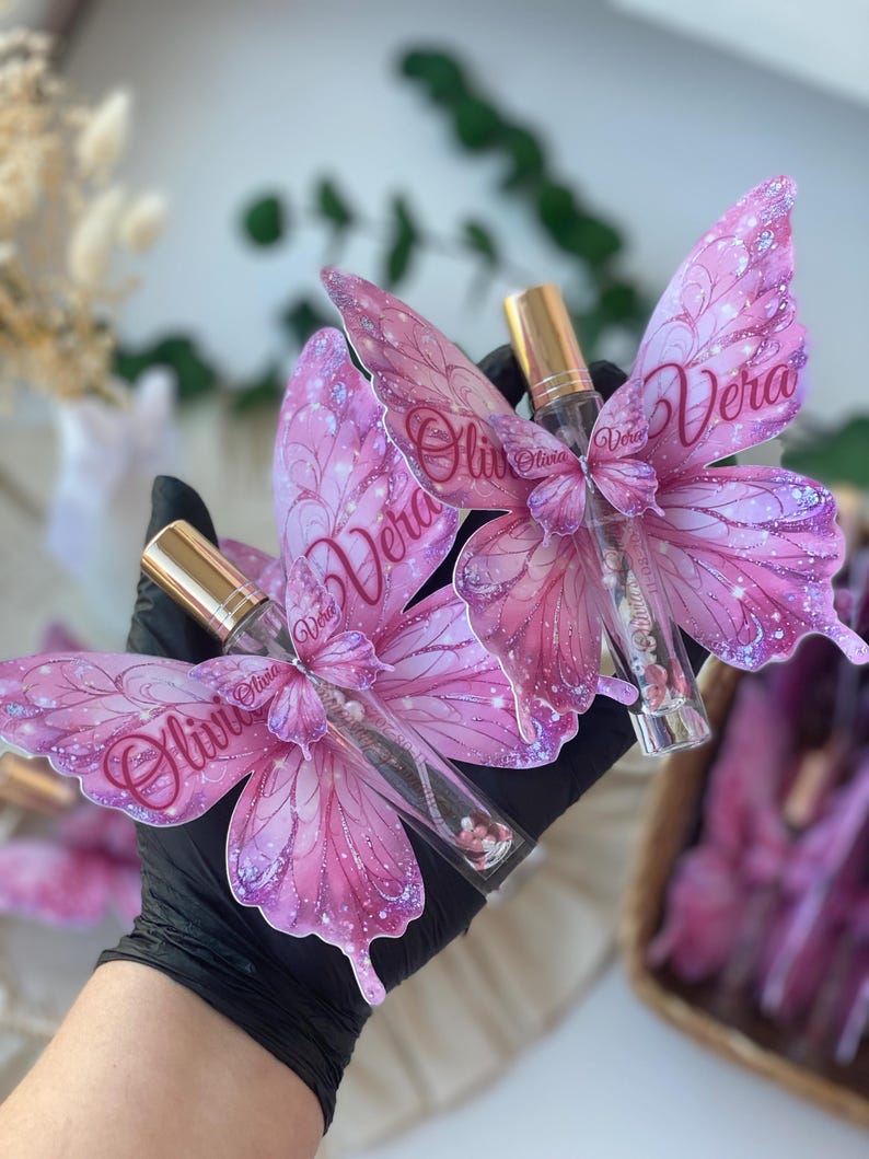 May include: Pink butterfly-shaped perfume bottles with gold caps. The butterfly wings are decorated with glitter and the names "Olivia" and "Vera". The bottles are held in a black gloved hand.