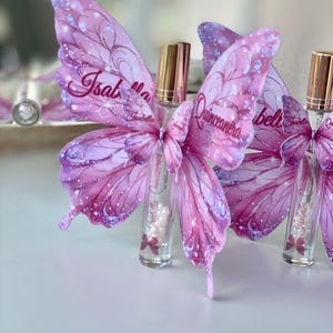 May include: A collection of clear glass perfume bottles with gold caps, each adorned with a pink butterfly wing design. The wings feature the name "Isabella Quincea&ntilde;era" in script, with glitter accents. The bottles are arranged on a white surface.