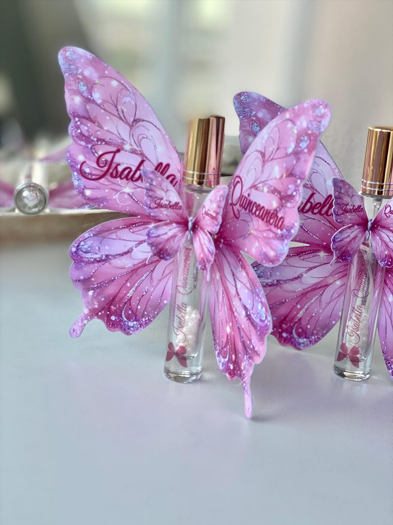 May include: A collection of clear glass perfume bottles with gold caps, each adorned with a pink butterfly wing design. The wings feature the name "Isabella Quincea&ntilde;era" in script, with glitter accents. The bottles are arranged on a white surface.