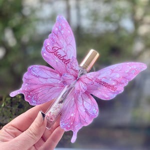 May include: A clear glass perfume bottle with a gold cap, adorned with a pink butterfly-shaped decoration. The butterfly wings are embellished with glitter and the name "Isabella Quincea&ntilde;era" is written on the wings.
