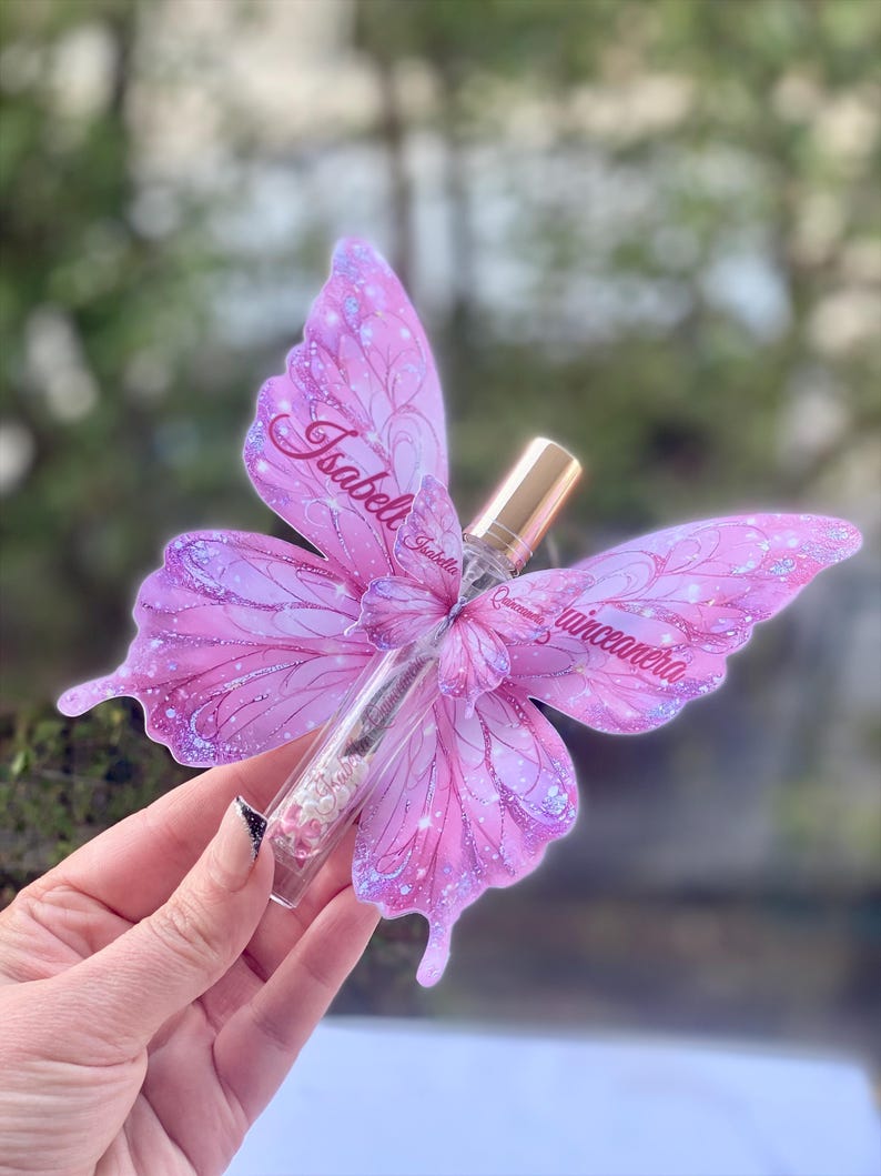 May include: A clear glass perfume bottle with a gold cap, adorned with a pink butterfly-shaped decoration. The butterfly wings are embellished with glitter and the name "Isabella Quincea&ntilde;era" is written on the wings.