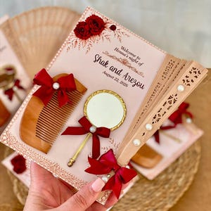 Può includere: Una carta rettangolare con il testo Welcome to Henna's night of Shak and Arezou August 22, 2026, decorata con rose rosse, un pettine in legno, un piccolo specchio rotondo con bordo dorato e un ventaglio in legno, accentuati da fiocchi rossi.