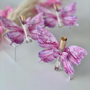 May include: Several butterfly-shaped favors with pink and purple wings, each adorned with the name "Isabella" and the word "Quincea&ntilde;era." The favors are arranged on a white surface, with a blurred background of pink flowers.