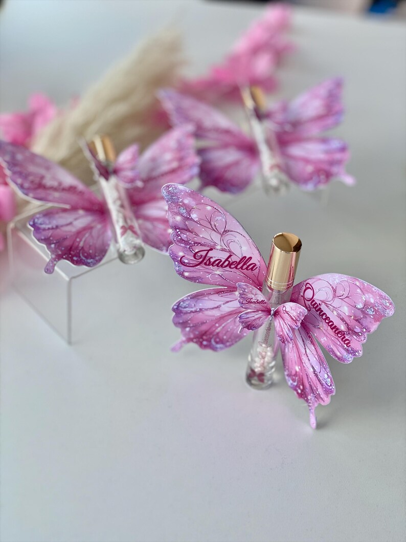 May include: Several butterfly-shaped favors with pink and purple wings, each adorned with the name "Isabella" and the word "Quincea&ntilde;era." The favors are arranged on a white surface, with a blurred background of pink flowers.