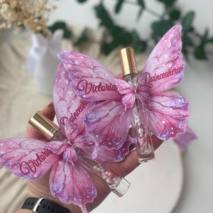 May include: Two perfume bottles with butterfly-shaped wings. The wings are pink with glitter accents and the name "Victoria" is printed on them. The bottles have gold caps and are filled with clear liquid and small pearls.
