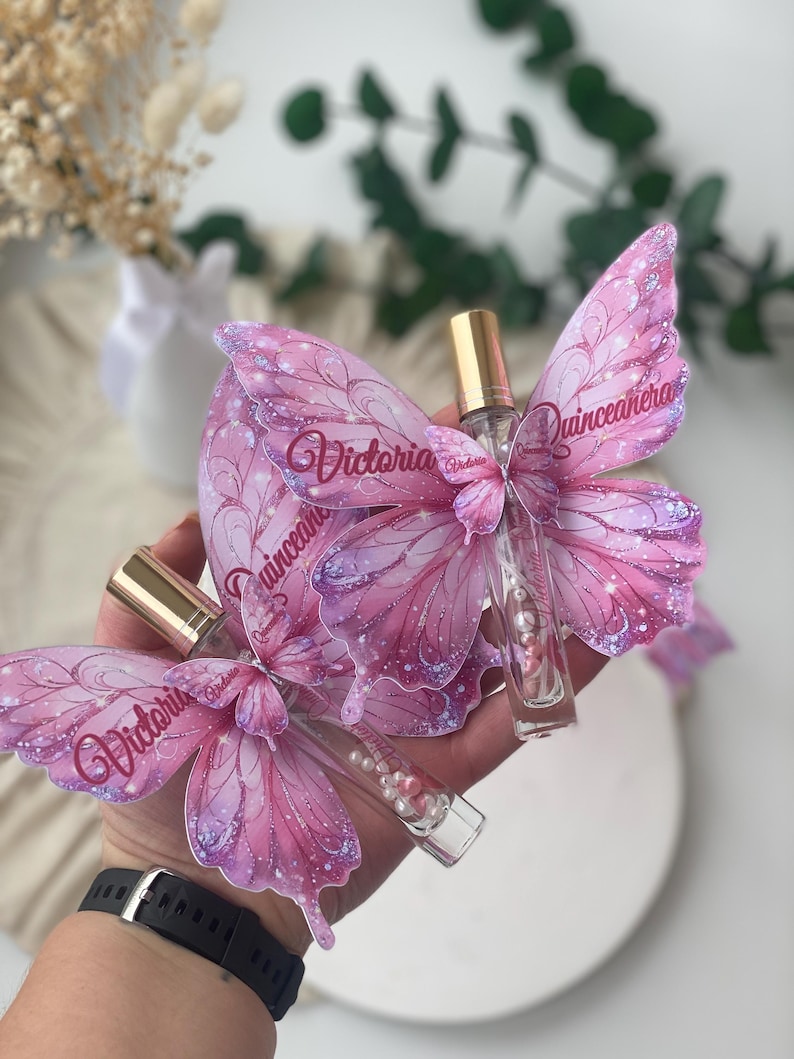 May include: Two perfume bottles with butterfly-shaped wings. The wings are pink with glitter accents and the name "Victoria" is printed on them. The bottles have gold caps and are filled with clear liquid and small pearls.