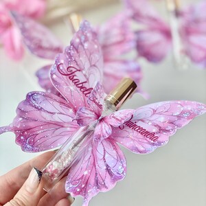 May include: A clear glass vial filled with pink beads and a gold cap, nestled within a pink butterfly-shaped decoration. The butterfly wings are adorned with glitter and the name "Isabell" and "Quinceanera".