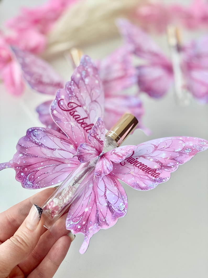 May include: A clear glass vial filled with pink beads and a gold cap, nestled within a pink butterfly-shaped decoration. The butterfly wings are adorned with glitter and the name "Isabell" and "Quinceanera".