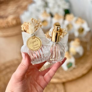 A hand holds two small, clear, ribbed glass perfume bottles with gold tops, white bows, dried flowers, and a gold tag reading "Kajal's Bridal Shower."