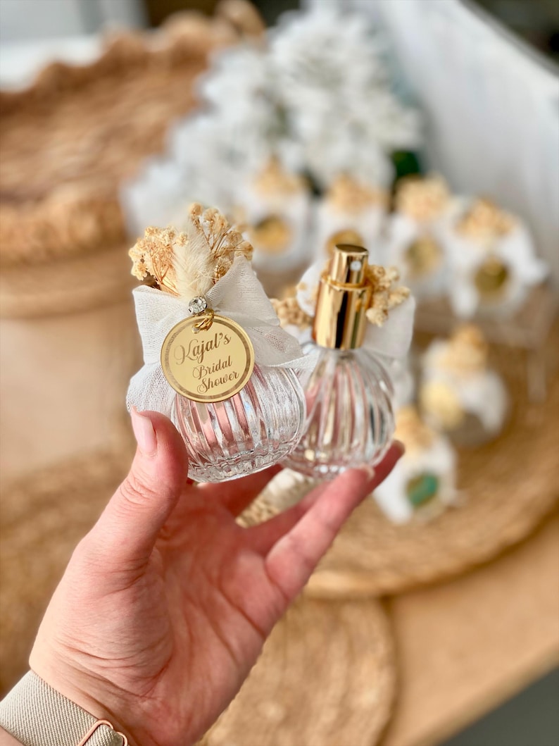 A hand holds two small, clear, ribbed glass perfume bottles with gold tops, white bows, dried flowers, and a gold tag reading "Kajal's Bridal Shower."