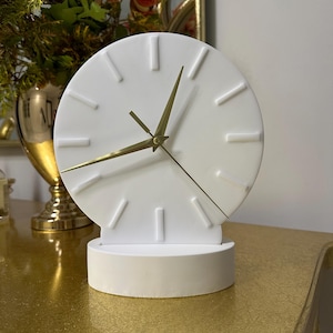 Peut inclure: Une horloge blanche minimaliste avec des aiguilles et des marqueurs d'heure dorés. L'horloge a un cadran circulaire et repose sur une base blanche assortie. Le design de l'horloge est moderne et élégant, adapté à la décoration intérieure.