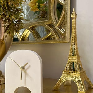 May include: A gold-colored Eiffel Tower model and a white arched clock with gold hands sit on a gold surface. A gold-framed mirror and a vase with greenery are in the background. The clock has a cut-out at the bottom.