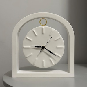Peut inclure: Une horloge de bureau blanche et minimaliste avec un cadran circulaire et des aiguilles noires. L'horloge est encadrée dans un cadre rectangulaire blanc avec un sommet en arc et un anneau doré. Le cadran de l'horloge comporte des repères horaires linéaires en relief.