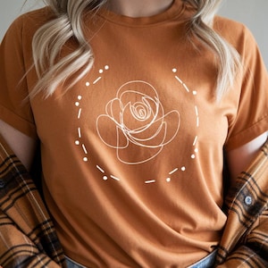 Puede incluir: Una camiseta de manga corta de color óxido con un dibujo lineal blanco de una rosa dentro de un círculo punteado. La camiseta se combina con vaqueros azules y una camisa de franela a cuadros marrón y negra.