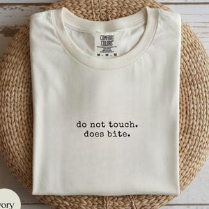 以下が含まれることがあります： 「do not touch. does bite.」の黒い文字が入ったアイボリー色のTシャツ。 シャツは折りたたまれ、織られた円形の表面に表示されています。 ブランドラベル「Comfort Colors」がネックラインに見えます。