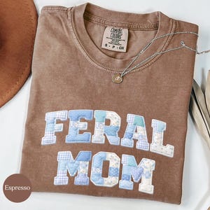 Op de afbeelding: Espresso-kleurig T-shirt met de tekst "FERAL MOM" in een patchwork-ontwerp van blauwe en witte stoffen. Een zilveren ketting met een kleine hanger ligt op het shirt. Het shirt is van het merk Comfort Colors, maat S-P-CH.