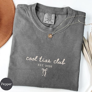 May include: A gray Comfort Colors t-shirt with the text "cool tias club" and "EST. 2026" printed in a light beige color. A silver necklace with a pendant is draped across the shirt. A brown hat is visible in the background.