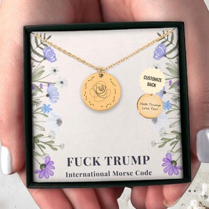 Könnte beinhalten: Eine goldfarbene Halskette mit zwei runden Anhängern. Der größere Anhänger zeigt ein Rosendesign und Morsecode. Der kleinere Anhänger sagt "Hate Trump Love You!" Auf der Schachtel steht "FUCK TRUMP International Morse Code."