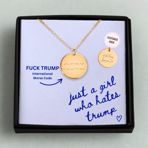 Puede incluir: Collar dorado con colgante redondo. El colgante muestra el texto "FUCK TRUMP" en código Morse internacional. El texto "just a girl who hates trump" está escrito en azul. Presentado en una caja negra.