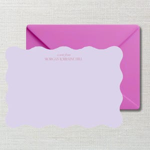 May include: A lilac-colored stationery set featuring a scalloped-edge note card and a magenta envelope. The note card has the text "a note from MORGAN LORRAINE HILL" printed in a soft pink hue. The stationery set is on a light beige background.
