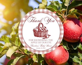 May include: A round sticker with a red and white gingham border and the words "Thank You" in elegant script. The center features a basket of apples. Text below reads "FOR SHOWERING THE APPLE OF OUR EYE, Karoline's Baby Shower, October 18, 2025."