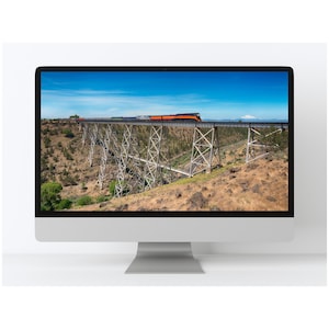 May include: A computer monitor displays a scenic image of a train crossing a high bridge. The train is orange and black, set against a blue sky. The bridge is made of metal and spans a canyon with dry, brown vegetation.