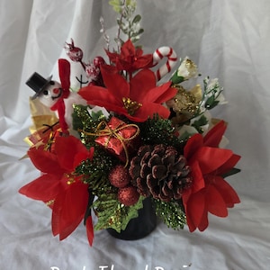May include: A festive floral arrangement in a black container featuring red poinsettia flowers, a pinecone, a small snowman figure, a red gift box, and a candy cane. The text Den's Floral Designs is printed at the bottom of the image.