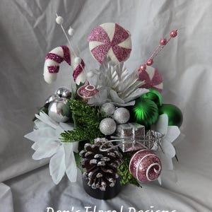 May include: A festive floral arrangement in a black container featuring white poinsettias, green and silver baubles, a pinecone, a glittery pink candy cane, a round pink peppermint sweet, and a small silver gift box. Text reads: Den's Floral Designs.