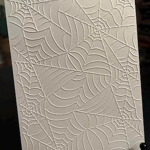 May include: A textured, off-white card featuring an embossed spiderweb design. The intricate pattern of webs is repeated across the surface, creating a visually interesting and thematic piece. The card is displayed on a black stand.