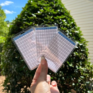 May include: Three clear plastic cards with white text and a blue header are held up against a green bush. The cards display a table of numbers, including percentages and measurements in cm.