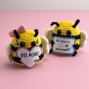 Op de afbeelding: Twee handgemaakte gehaakte bijenbeeldjes. Eén bij heeft een roze strik en een hartvormig bordje met de tekst "BEE MINE!". De andere bij houdt een bord vast met de tekst "BEE-lieve in yourself!". Beide bijen zijn geel met zwarte strepen en vleugels.