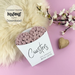 Pode incluir: Dois  coasters em forma de coração de crochê rosa em um suporte de papel branco com o texto "Coasters 100% Handmade with Love".