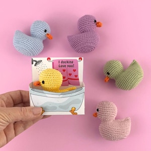 Può includere: Un set di cinque giocattoli di anatra all'uncinetto in vari colori, tra cui viola, azzurro, verde e rosa. Le anatre sono disposte su uno sfondo rosa. Un'anatra è in una vasca da bagno su una carta che dice "Ti amo da morire!"
