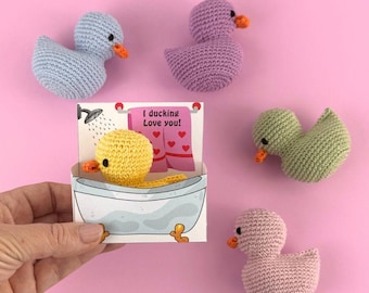 Puede incluir: Un conjunto de cinco juguetes de pato de crochet en varios colores, incluyendo morado, azul claro, verde y rosa. Los patos están dispuestos sobre un fondo rosa. Un pato está en una bañera en una tarjeta que dice "¡Te quiero mucho!"