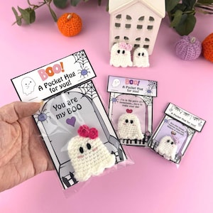 Könnte beinhalten: Handgefertigte gehäkelte Geister-Taschenumarmungen, jeweils verpackt mit einer Karte mit der Aufschrift "Boo! A Pocket Hug for you!" und anderen Sätzen. Die Geister sind weiß mit rosa Schleifen und schwarzen Augen. Dekorative Kürbisse und ein kleines Haus sind im Hintergrund.