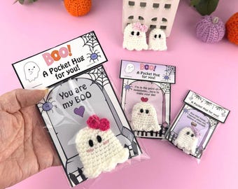 Op de afbeelding: Een verzameling handgemaakte gehaakte spookzakknuffels, elk verpakt met een kaart met de tekst "Boo! A Pocket Hug for you!" en andere zinnen. De spoken zijn wit met roze strikken en zwarte ogen. Decoratieve pompoenen en een klein huisje staan op de achtergrond.