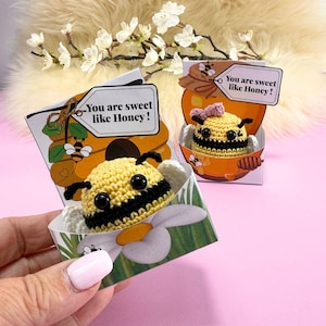 Puede incluir: Figuras de abeja de ganchillo en cajas decorativas. Las abejas son amarillas y negras, con antenas negras y ojos grandes negros. Las cajas presentan un diseño de panal y tarro de miel, con el texto "You are sweet like Honey!"
