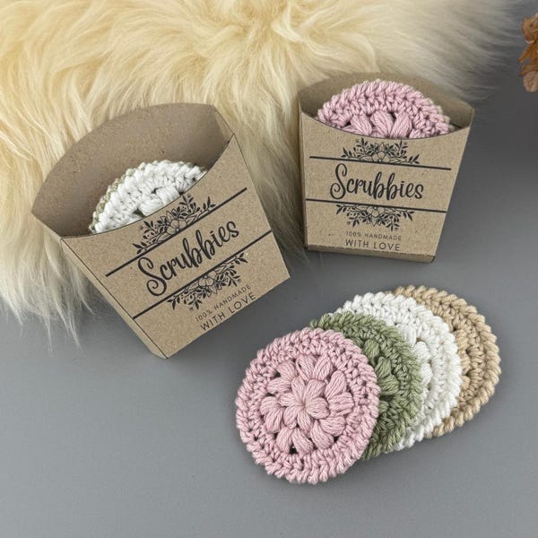 May include: A collection of handmade crochet scrubbies in various colors, including pink, green, white, and beige. The scrubbies are packaged in small, brown cardboard boxes labeled "Scrubbies" with floral accents and the text "100% Handmade With Love."