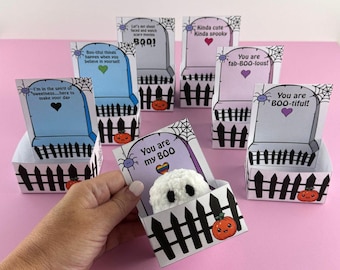 Puede incluir: Una colección de cajas de regalo con temática de Halloween con forma de lápidas, cada una con un mensaje y diseño únicos. Las cajas presentan un diseño de valla de piquetes negros, telarañas e ilustraciones de calabazas. Una caja contiene un pequeño fantasma blanco.